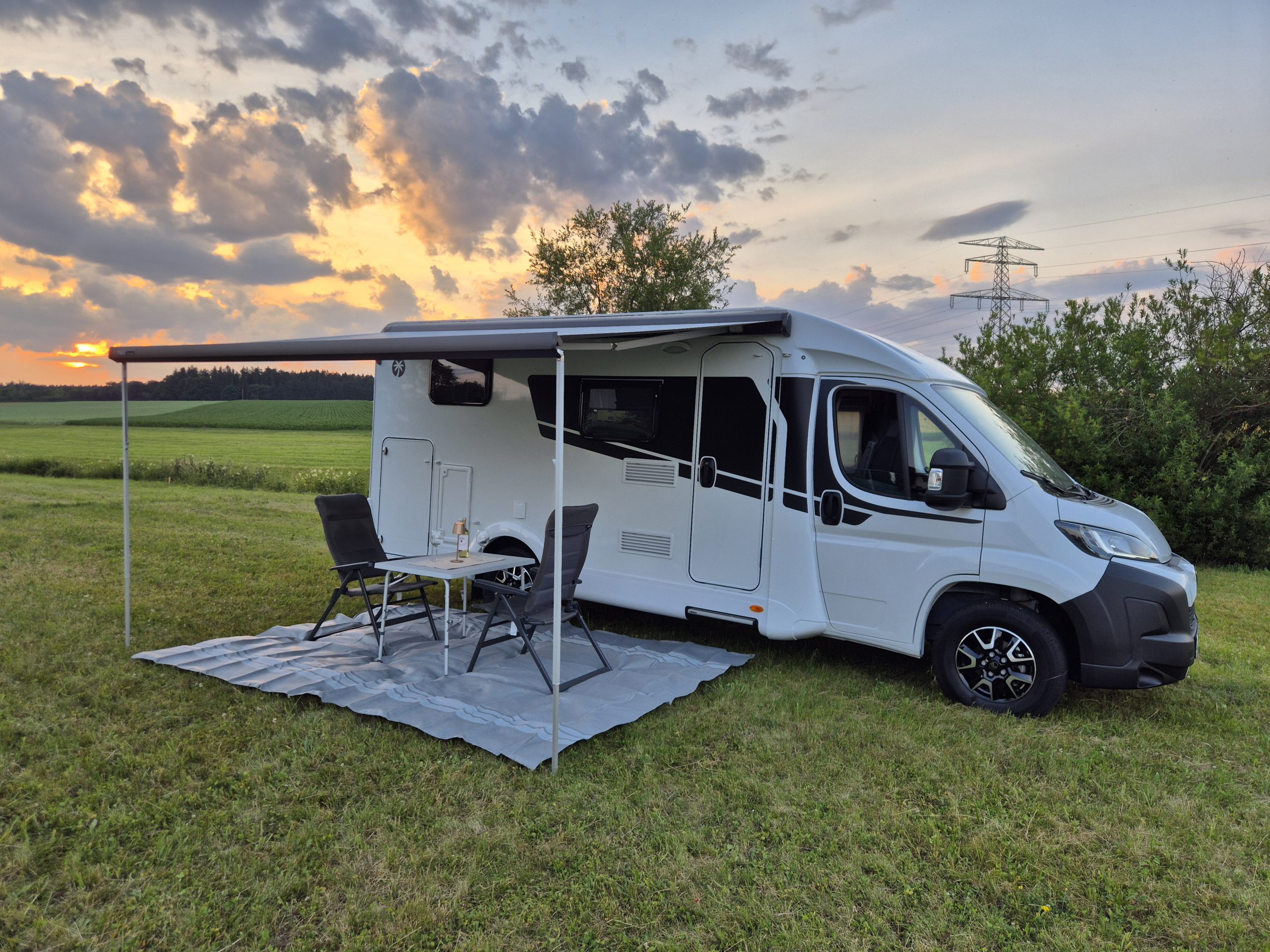 Wohnmobil mit ausgefahrener Markise, Sitzgruppe und Tisch bei Sonnenuntergang auf einer Wiese